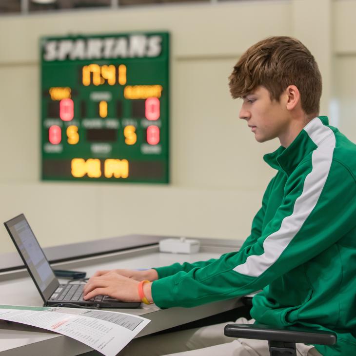 Sport Management student working on a laptop with a scoreboard in the background