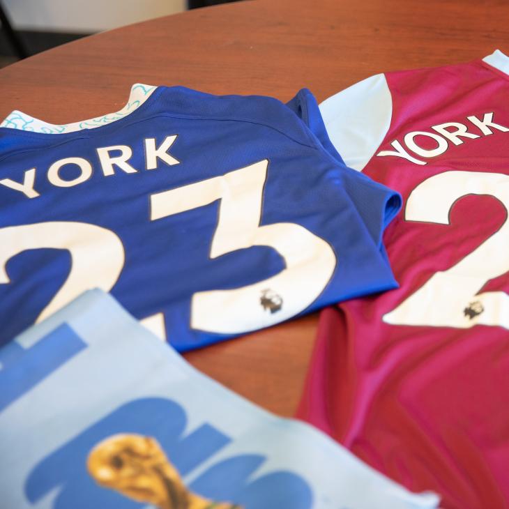 A blue jersey with number 23 and the name "York" on the back and a red jersey with the number 20 and name "York College" are laid out on a table in an office.
