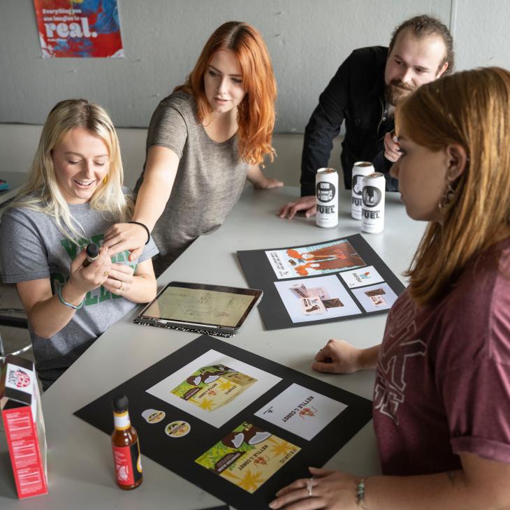 A group of four students gather around a table covered with mock-ups and sketches in the graphic design studio.