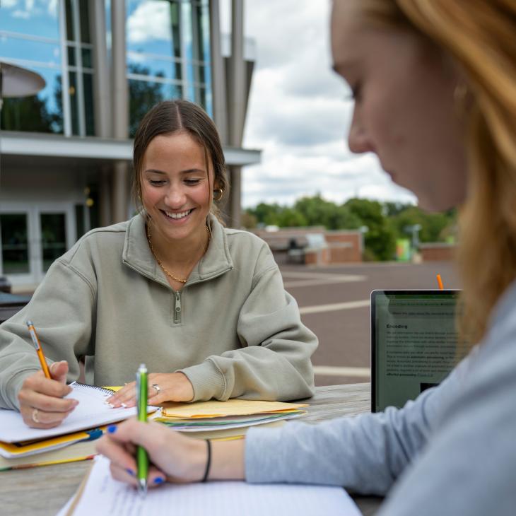 Two students smile while writing in notebooks outdoors at a picnic table on the campus quad.