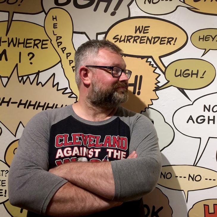 Artist Matthew Borgen poses, arms crossed, in front of a wall of his artwork — a series of comic-style speech bubbles