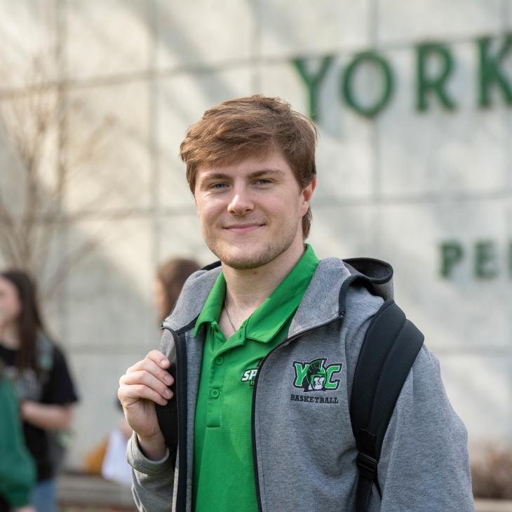 A student smiles at the camera on campus, dressed in a York College green polo and jacket, carrying a backpack.
