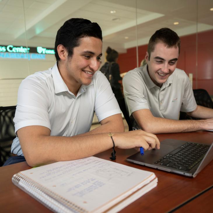 Two students look at a laptop in a classroom. The NASDAQ ticker is visible on the lobby wall outside the room's glass wall.