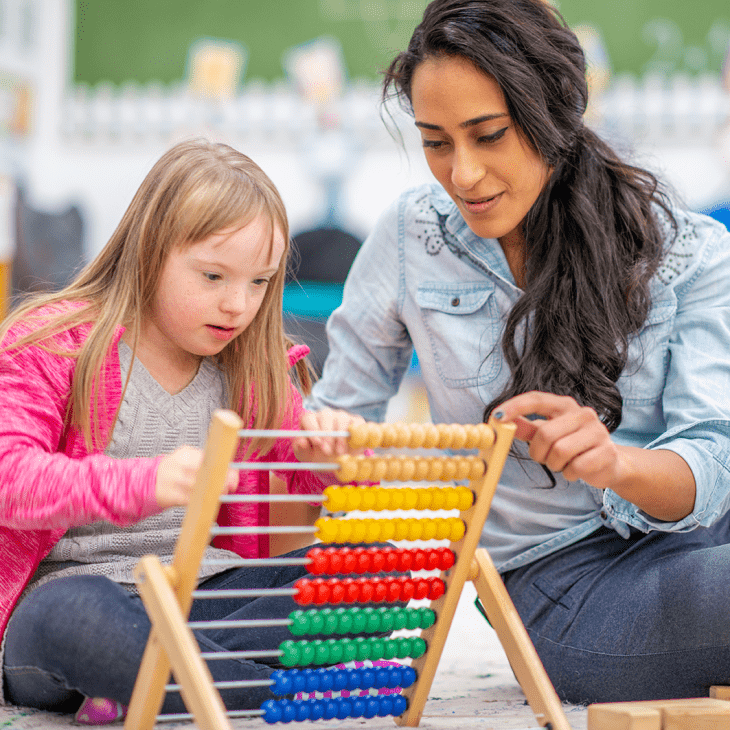 An elementary school teacher and student sit on the classroom floor, working with an abacus.