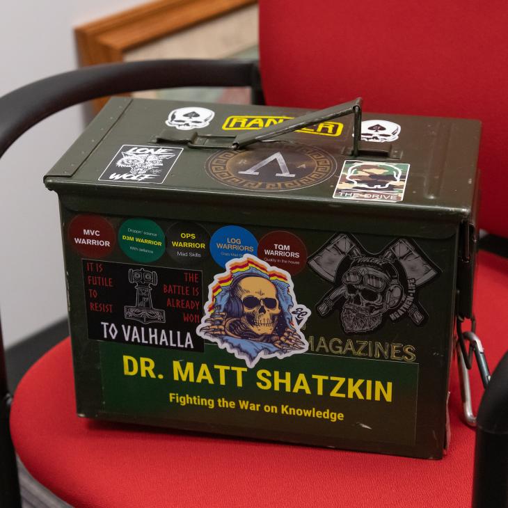 An ammunition can sits on a red office chair. It is covered in stickers and text on the side reads "Dr. Matt Shatzkin - Fighting the War on Knowledge"