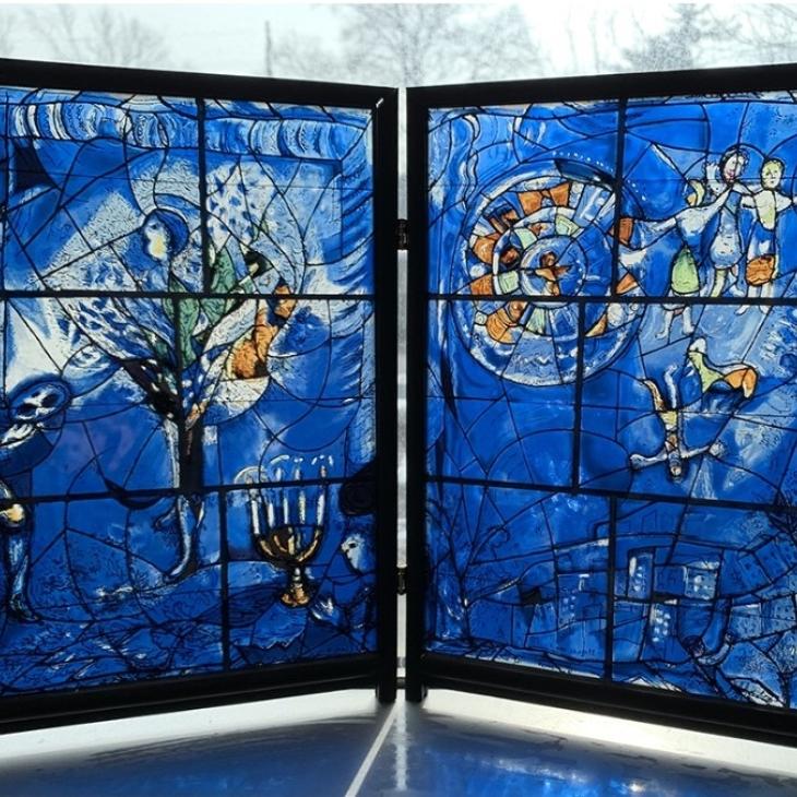 A reproduction of the third panel of Marc Chagall’s stained-glass window triptych, America Windows, at the Art Institute of Chicago. The glass is stained with a bright, deep blue base and includes burnt orange accented figures.