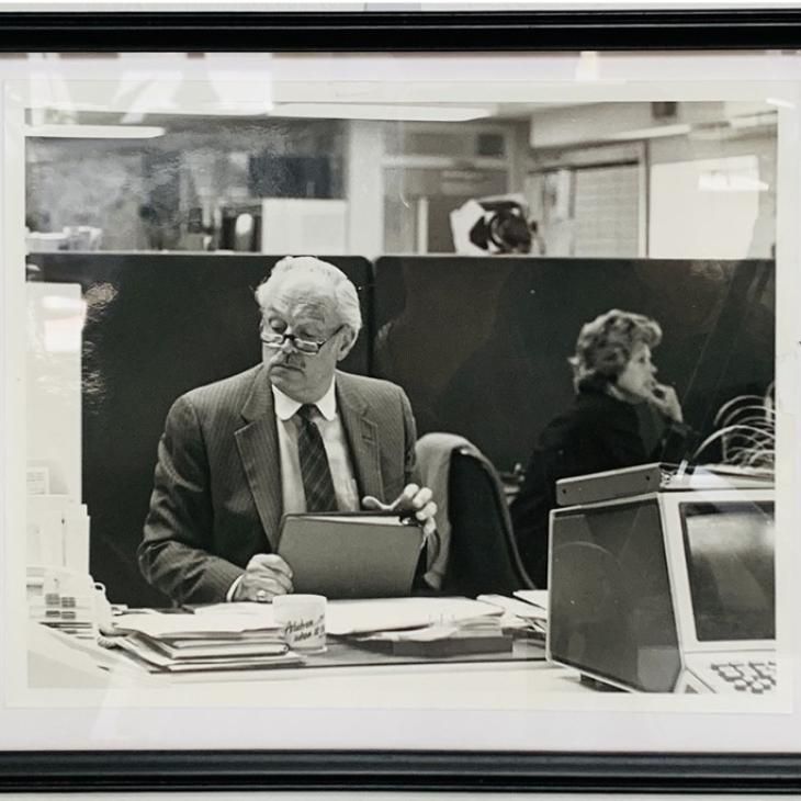 Photo of Tom Miller, Professor Rodger's father, seated at a desk, covered with papers and folders, holding a three-ring binder in his right hand.