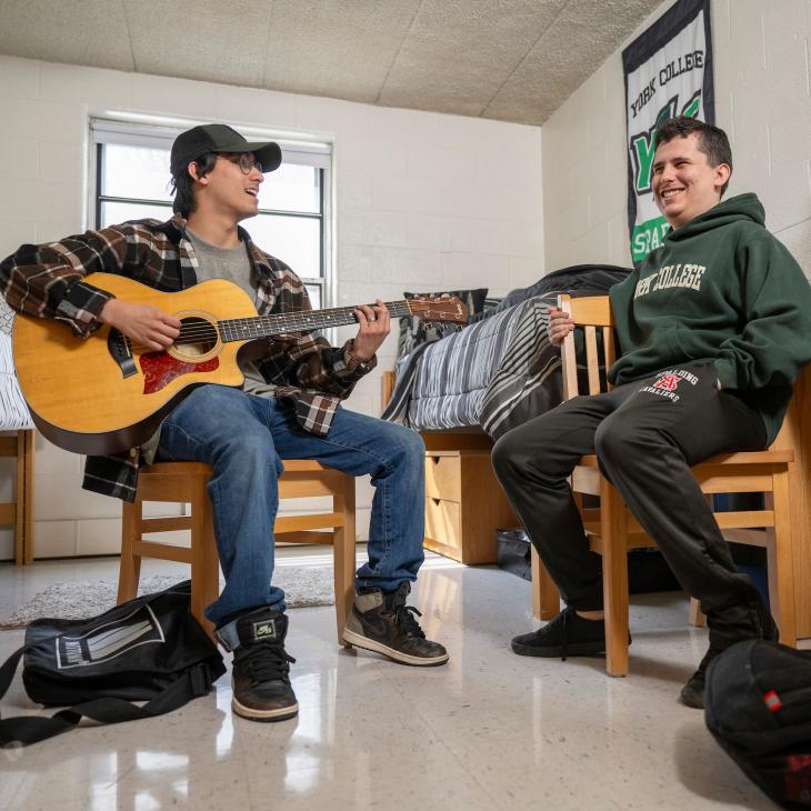 Students, one playing a guitar, in residence hall room. 