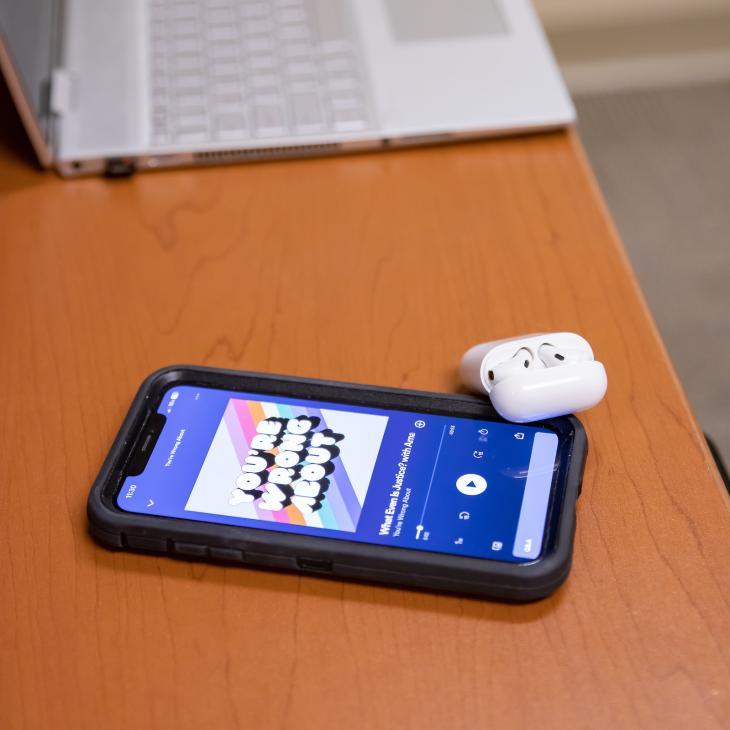 Airpods and a phone with a podcast app on screen sit on a desk.