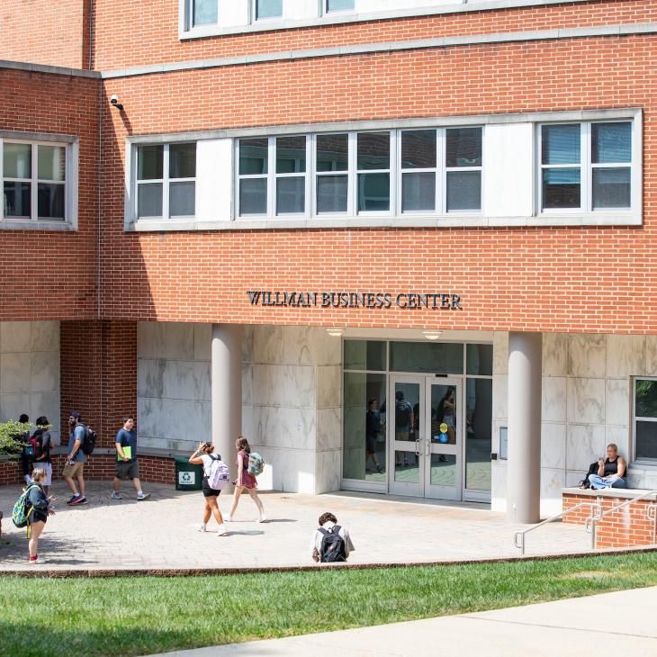 Students walk past a large brick building with signage identifying it as the Willman Business Center.