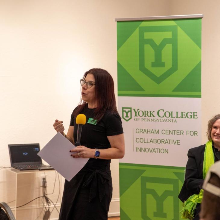 Tamara Sniad, Ph.D., speaks into a microphone while holding notes in front of a banner for the Graham Center for Collaborative Innovation at York College of Pennsylvania.