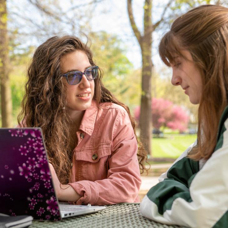 Two students sitting at an outdoor table, one is taking notes on a laptop.