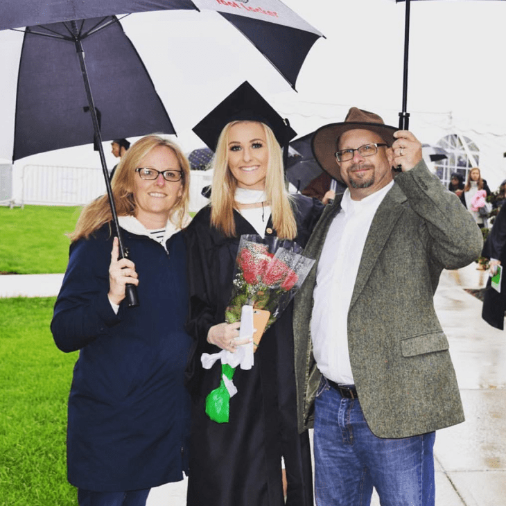 College graduate in cap and gown holds flowers and smiles with parents on either side holding umbrellas after a rainy commencement ceremony.
