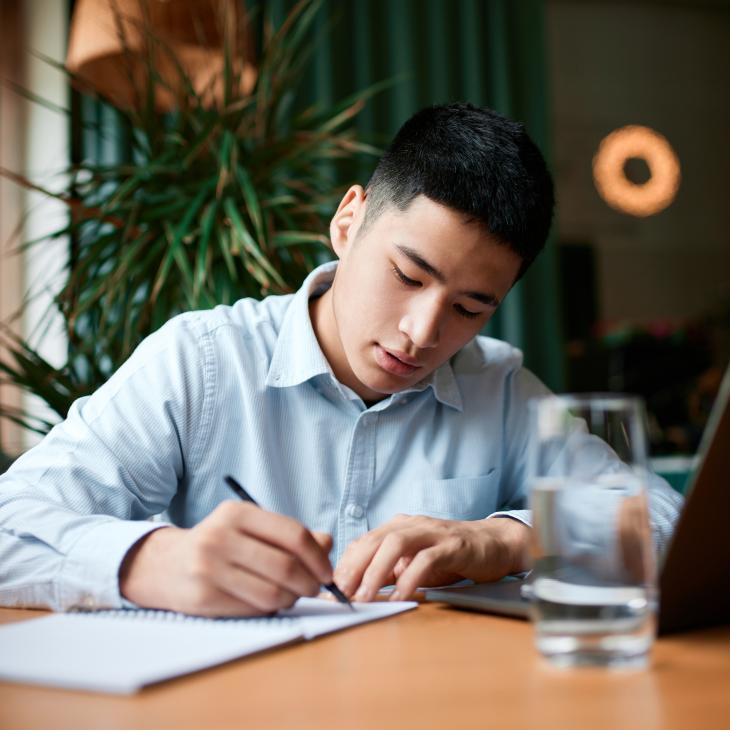 Person writing in a notebook with a laptop facing them.
