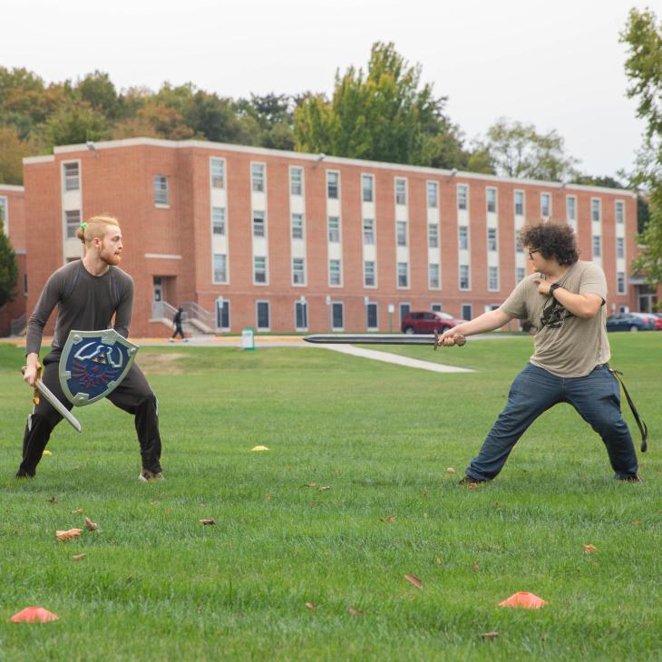 Two college students engaged in live action role play on a grassy lawn on campus.