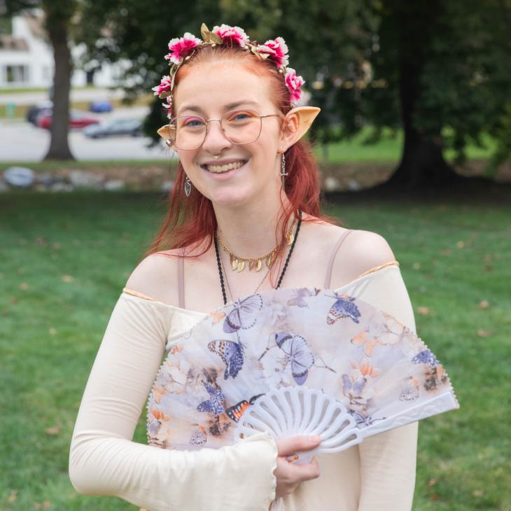 A student in a costume with pointy ears smiles at the camera while displaying a paper fan in an outdoor setting.