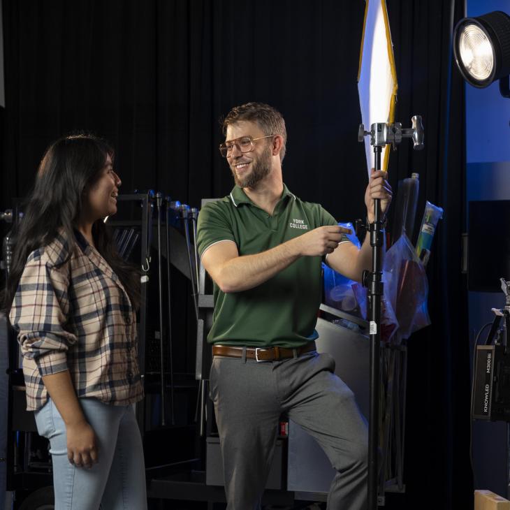 A male and female student stand together with equipment in a tv studio with lights behind them.