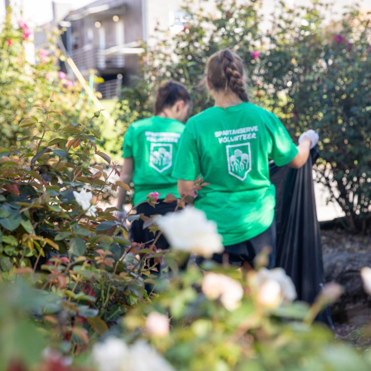 Two individuals wearing green tee shirts with white text reading "Spartan Volunteer Network" appear to be walking away from the camera while holding a black trash bag. They are outdoors with green, flowering shrubs visible around them.