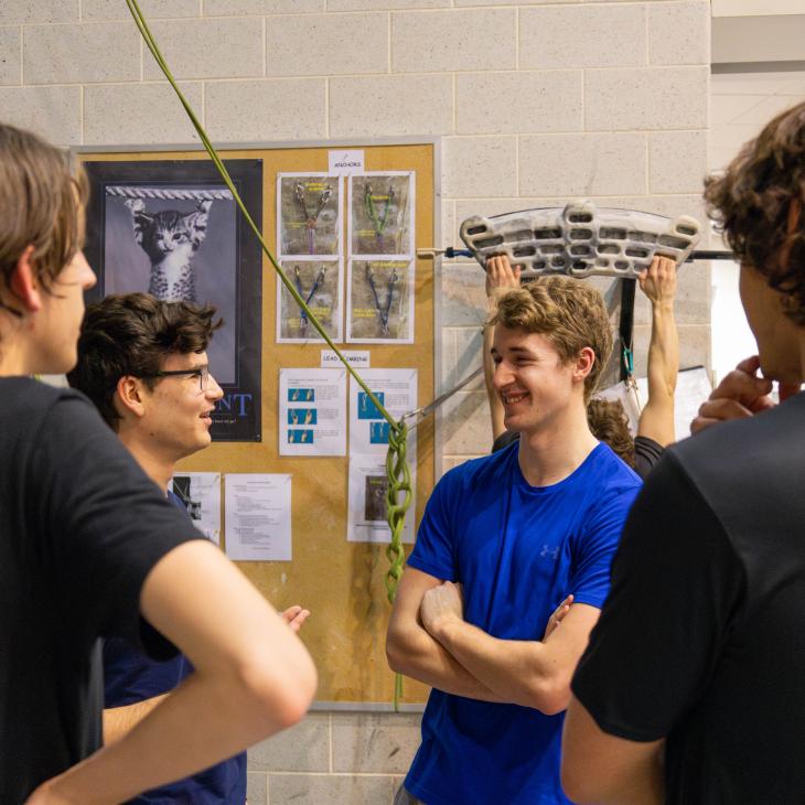A group of students engaged in casual conversation. A bulletin board with rock climbing related items is visible behind them.