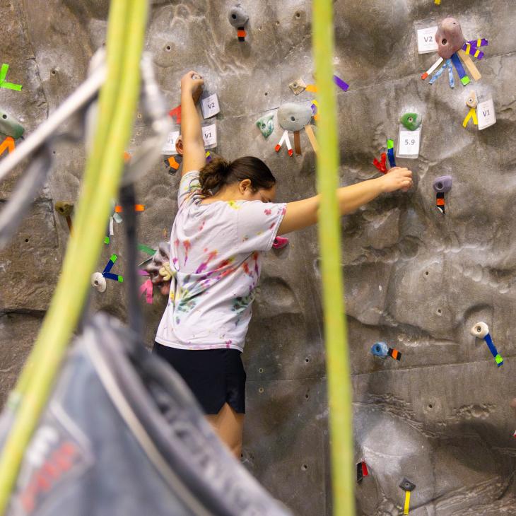 A rock climber begins an ascent on an indoor rock wall. A chalk bag and climbing ropes are visible in the foreground.