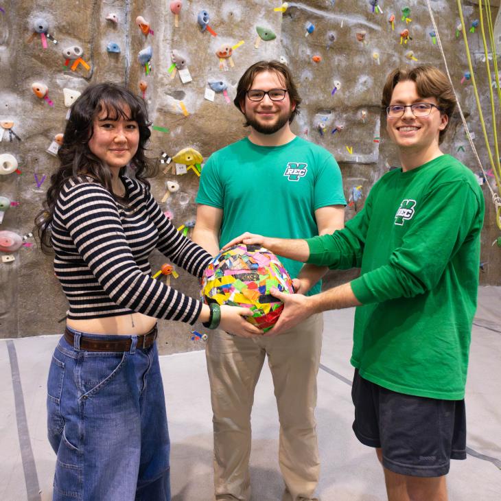 Three people standing, smiling at the camera while holding a colorful ball of tape between them.