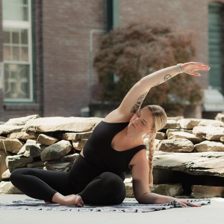 Kirsten Blair-Johnson ’18 practices a seated side stretch on a yoga mat outdoors near a stone wall and brick building.