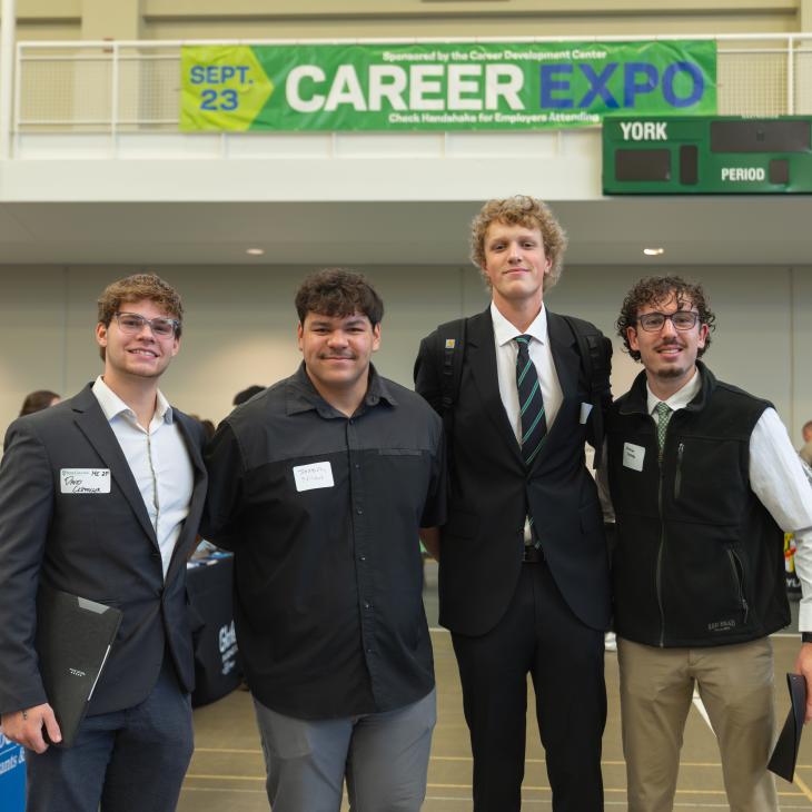 Four students stand together under a career expo banner in dress clothes.