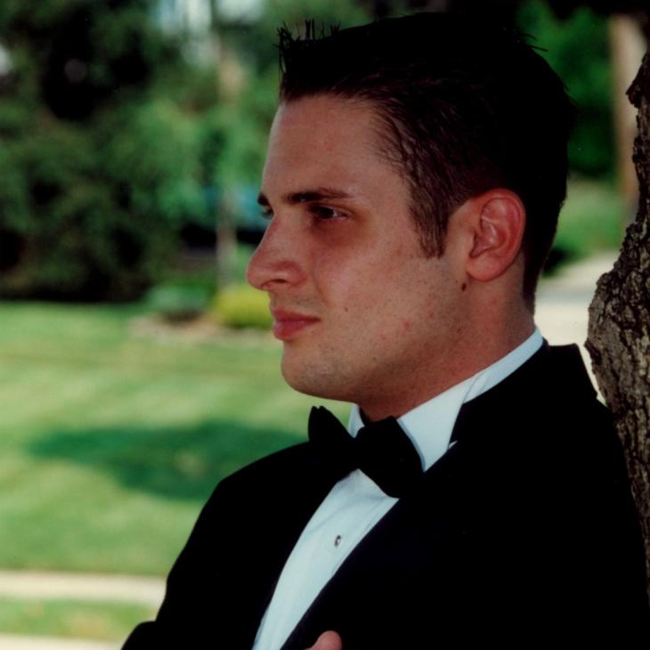 A man in a black tuxedo and bow tie stands outdoors with his arms crossed, leaning against a tree and looking to the side.