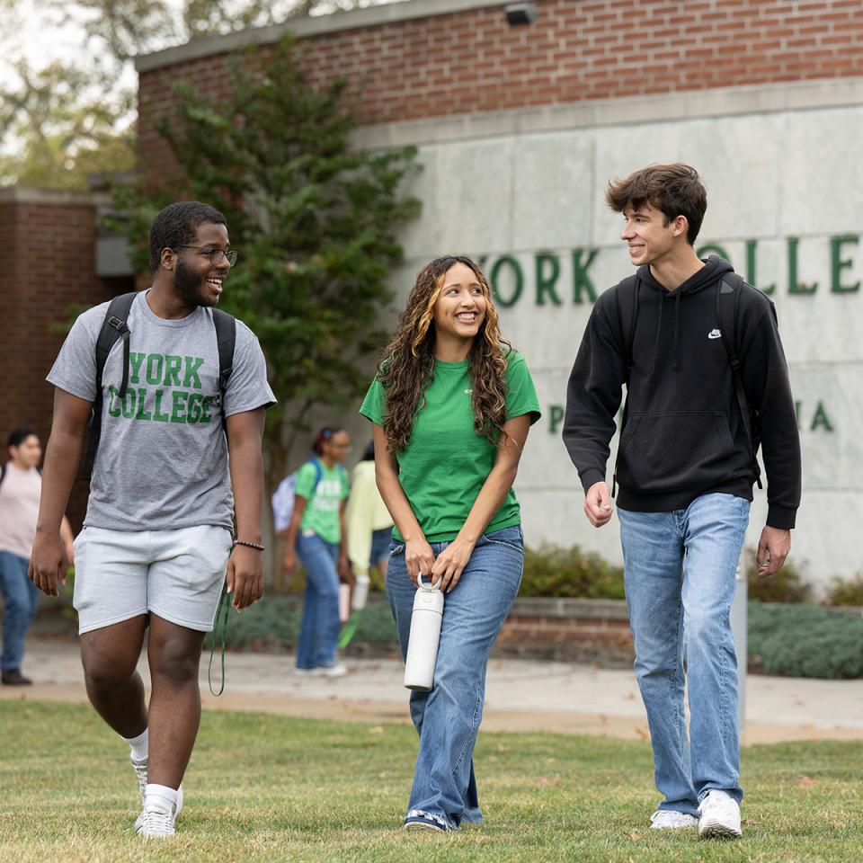 Three students walk across the front lawn with the York College sign behind them