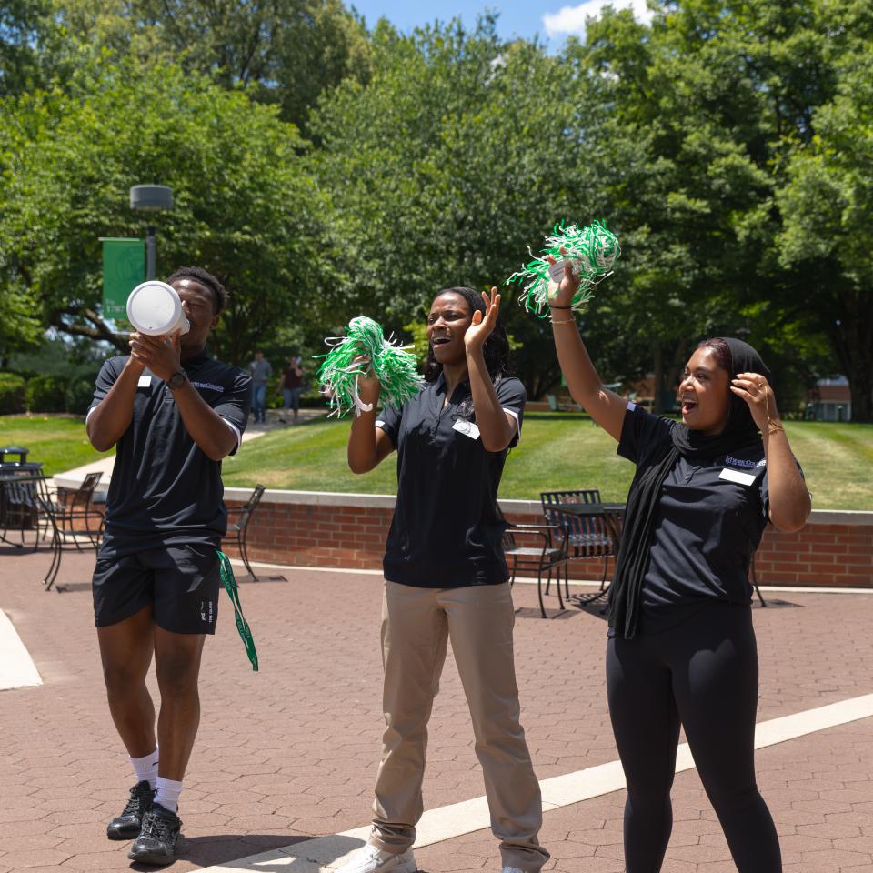 Student ambassadors stand outdoors on a sunny campus, cheering and welcoming visitors.