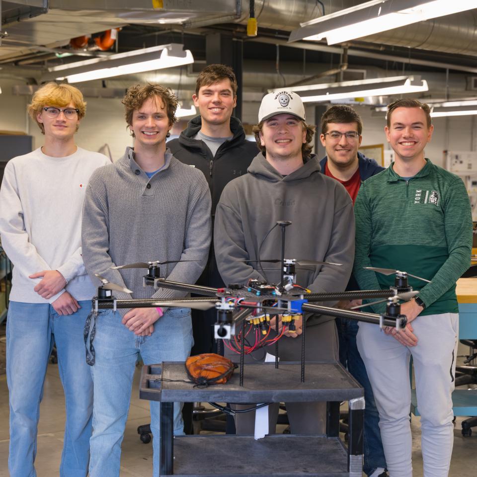 Six engineering students stand together in a laboratory behind a large quadcopter drone prototype displayed on a workbench.