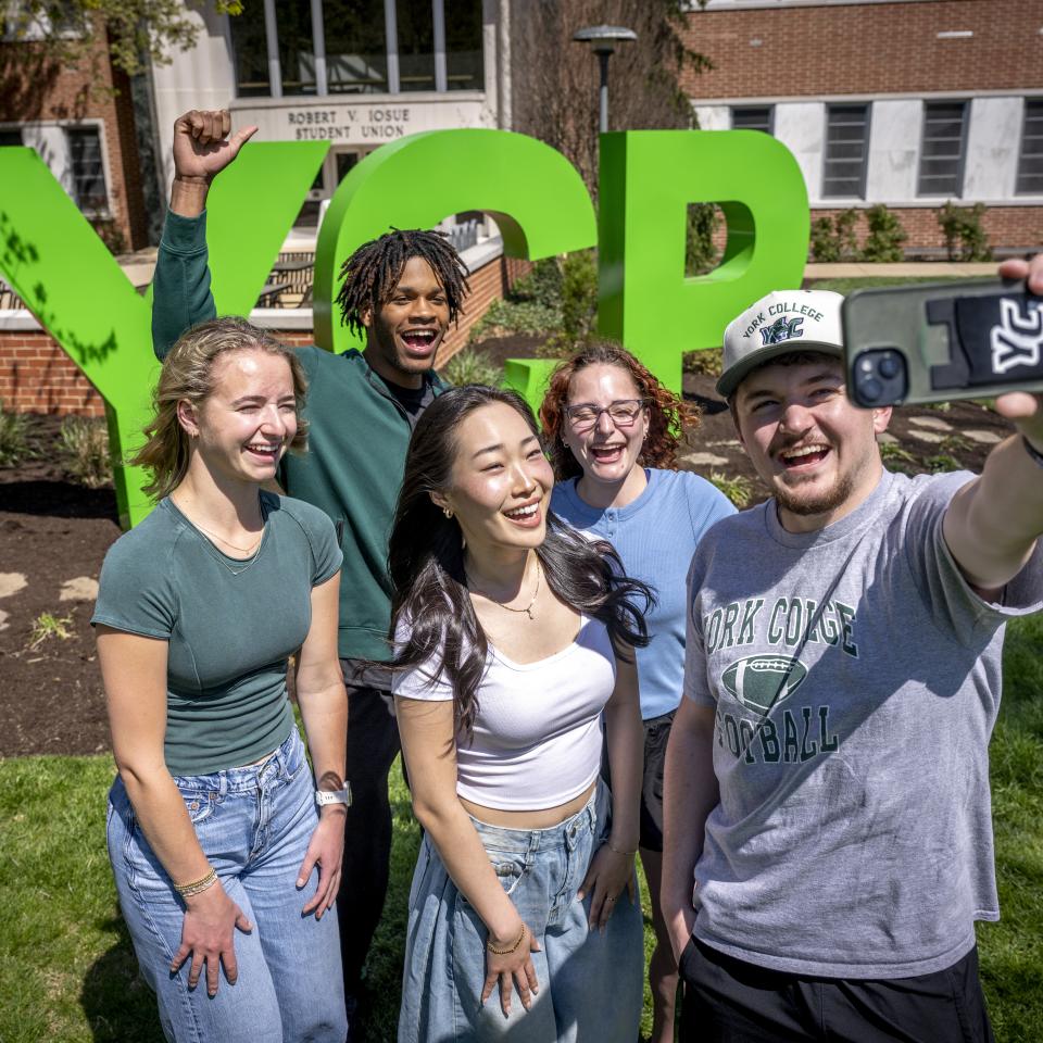 Five students smile while taking a selfie in front of large green "YCP" letters on campus, with the student union building visible in the background.