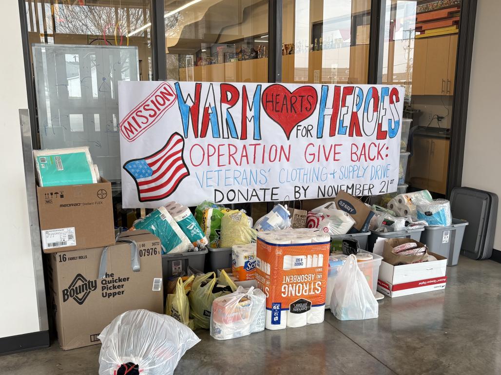 Assorted donated items are gathered in front of a hand-painted sign that reads, "Veterans' Clothing + Supply Drive."