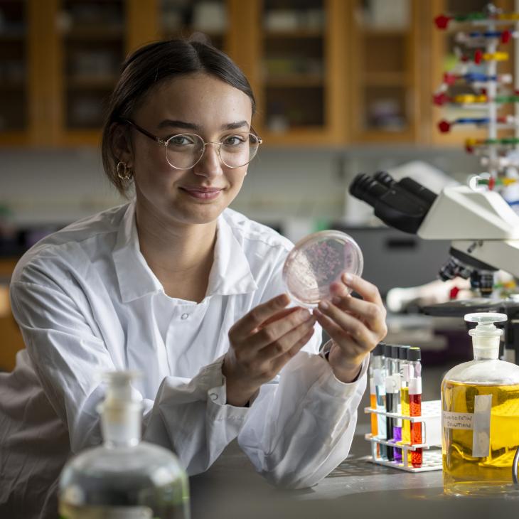A student in a science lab holds up a sample next to a microscope.