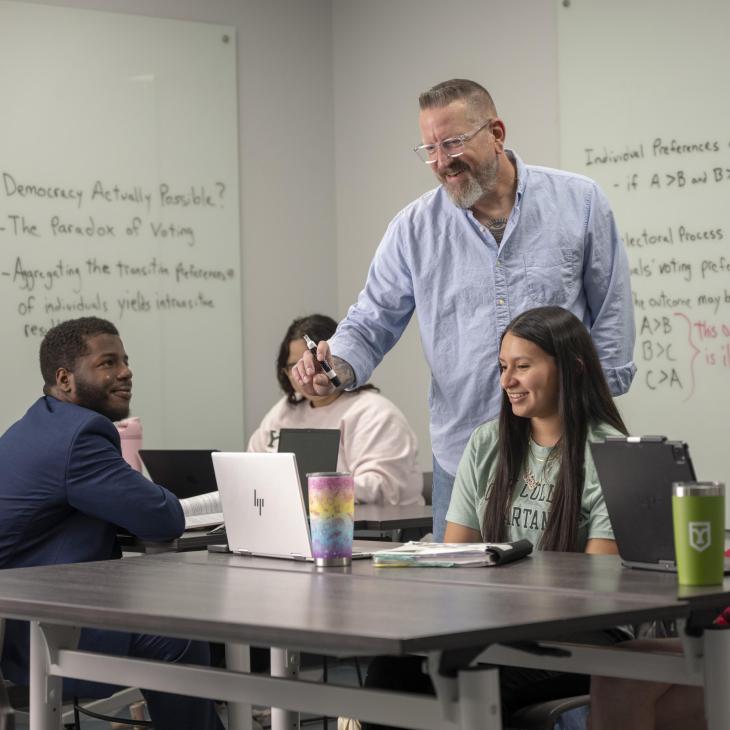 A teacher points to a computer in a classroom. He is surrounded by students and has a whiteboard behind him.